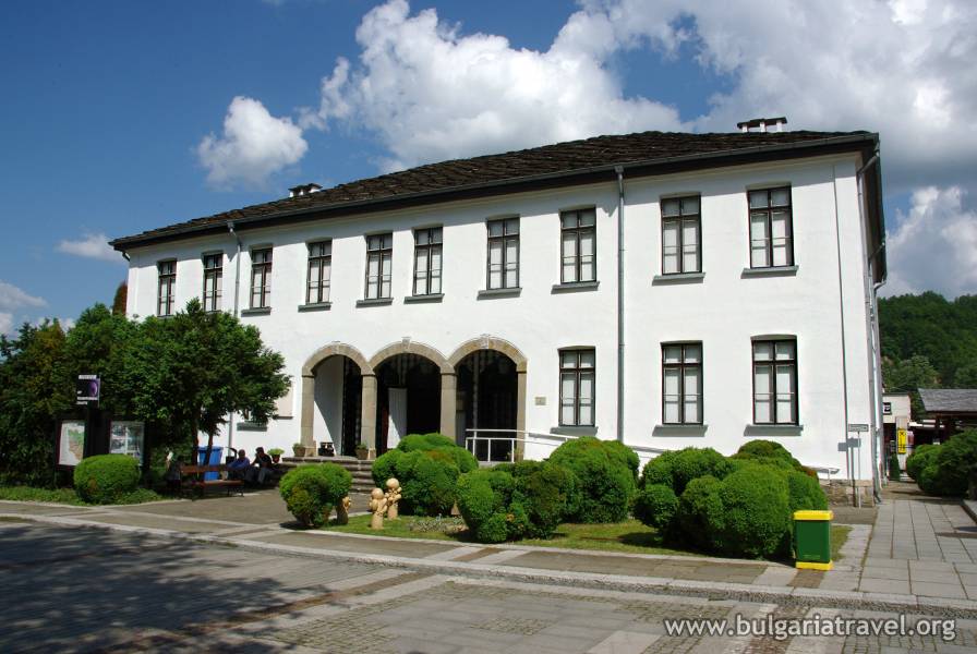 A spacious white building surrounded by lush greenery, featuring numerous trees and bushes.