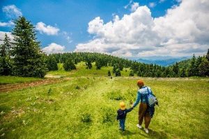 Жена и малко дете, държащи се за ръце, вървят през зелена поляна в планината под синьо небе с облаци.