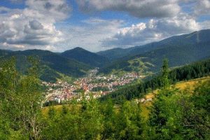 Панорамна гледка към малък планински град, заобиколен от гъсти гори и хълмове при ясно небе.