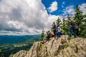 Група туристи, стоящи върху скален връх с изглед към планинска долина под облачно небе.