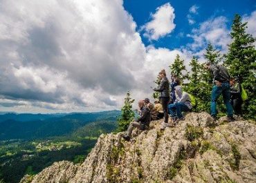 Група туристи, стоящи върху скален връх с изглед към планинска долина под облачно небе.